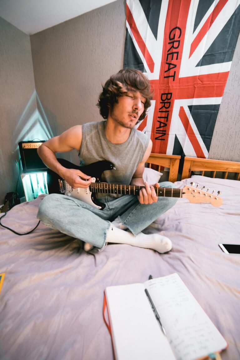 A young man plays an electric guitar on a bed, with a notebook nearby and a British flag in the background, creating a relaxed and creative atmosphere.