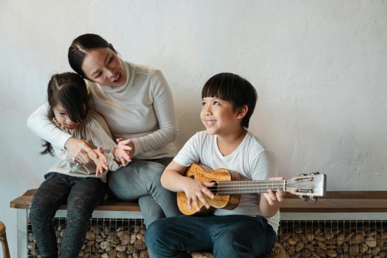 Dreamy ethnic brother in casual wear playing Hawaiian musical instrument while looking up and sitting near little sister and mother on wooden bench